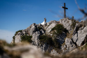 Hochzeit Bergshooting Christina und Michael - Brautpaar steht auf einem Berg in der Natur und küsst sich, das Gipfelkreuz sieht man im Bild rechts daneben.