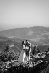 Hochzeit Bergshooting Christina und Michael - Brautpaar steht auf einem Berg in der Natur - Bräutigam und Braut schauen gemeinsam auf den Brautstrauß