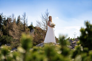 Hochzeit Bergshooting Christina und Michael - Braut steht auf einem Berg in der Natur und lächelt in die Kamera