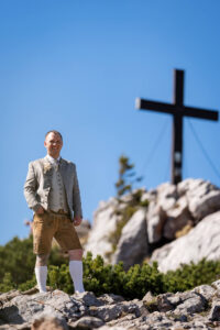 Hochzeit Bergshooting Christina und Michael - Bräutigam steht auf einem Berg in der Natur das Gipfelkreuz sieht man im Bild rechts daneben.