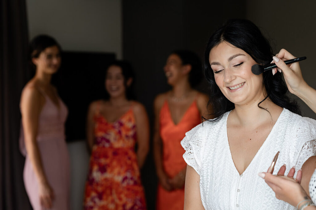 Hochzeit Julia und Stefan - Braut und Brautjungfern - Braut wird geschminkt und Brautjungfern stehen im Hintergrund - Braut lächelt in die Kamera