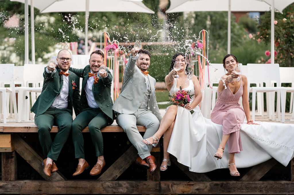 Hochzeit Julia und Stefan - Brautpaar und Beistände sitzen auf einem Steg und schütten Sekt aus Sektgläsern in die Höhe